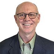 A headshot photo of Dr. Patrick Calhoun,  wearing a olive green sportsjacket and a dark blue button up shirt.
