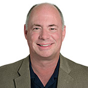 A headshot photo of Dr. Patrick Calhoun,  wearing a olive green sportsjacket and a dark blue button up shirt.